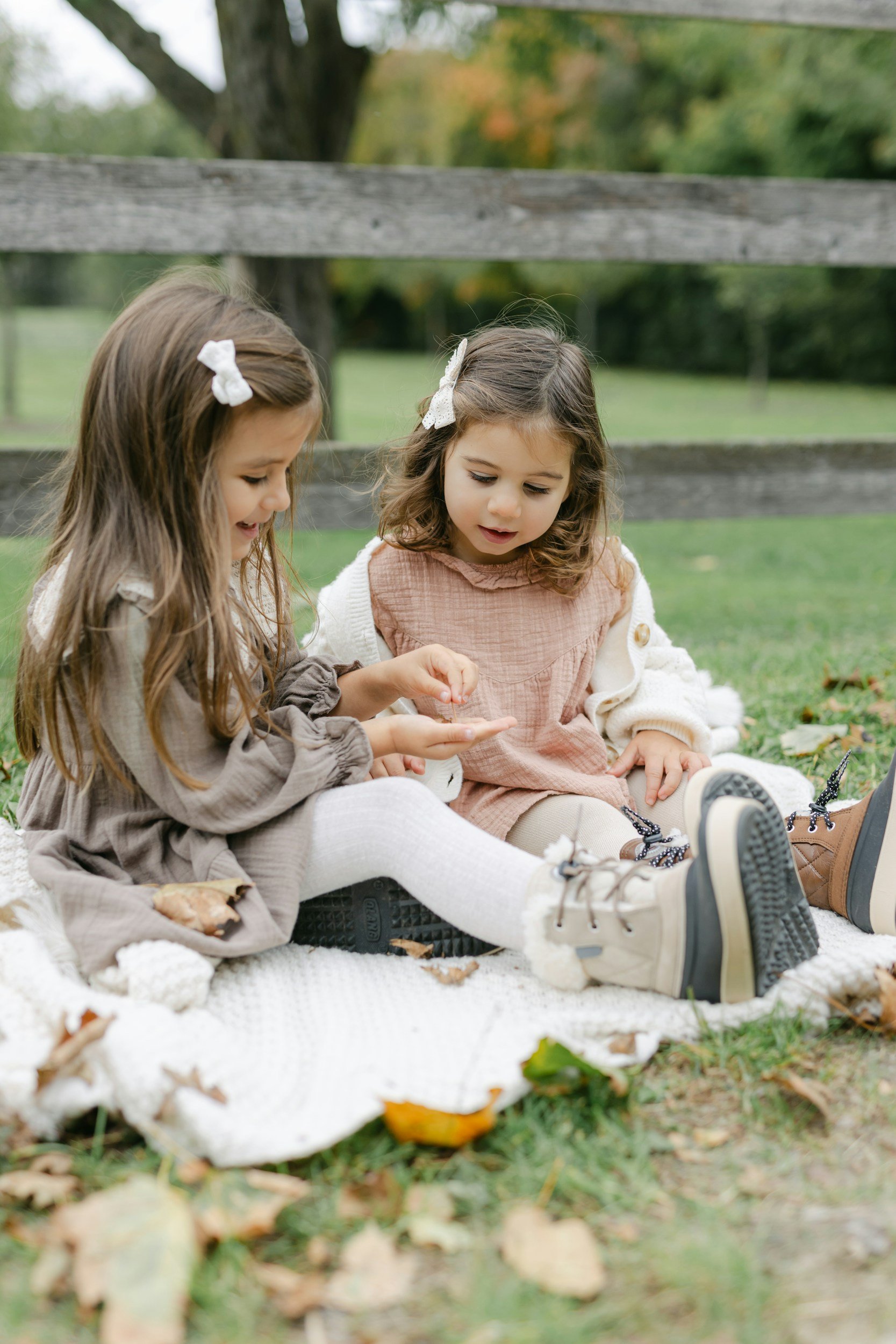 Two young girls sitting outdoors on a blanket, playing and examining leaves, with a wooden fence and trees in the background.