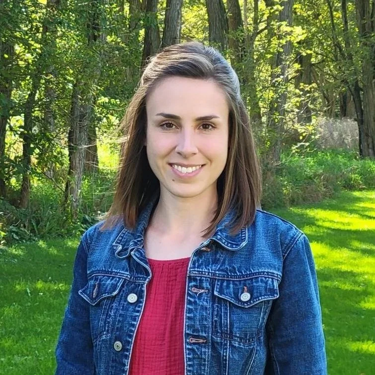 A young woman with shoulder-length brown hair, wearing a denim jacket and a red top, standing outdoors in a green, wooded area with sunlight filtering through trees.