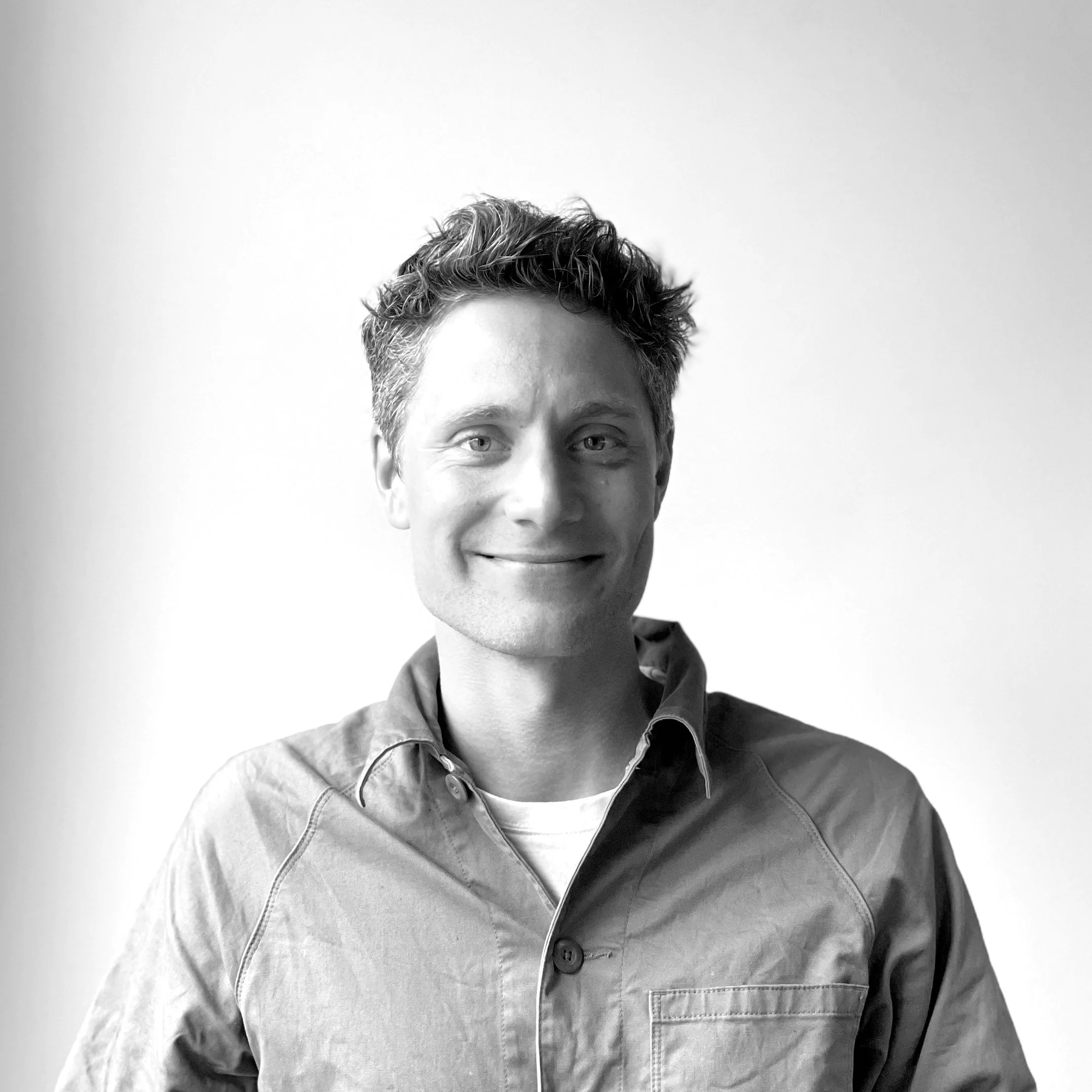 Black and white portrait of a smiling young man with curly hair, wearing a button-up shirt, against a plain light background.