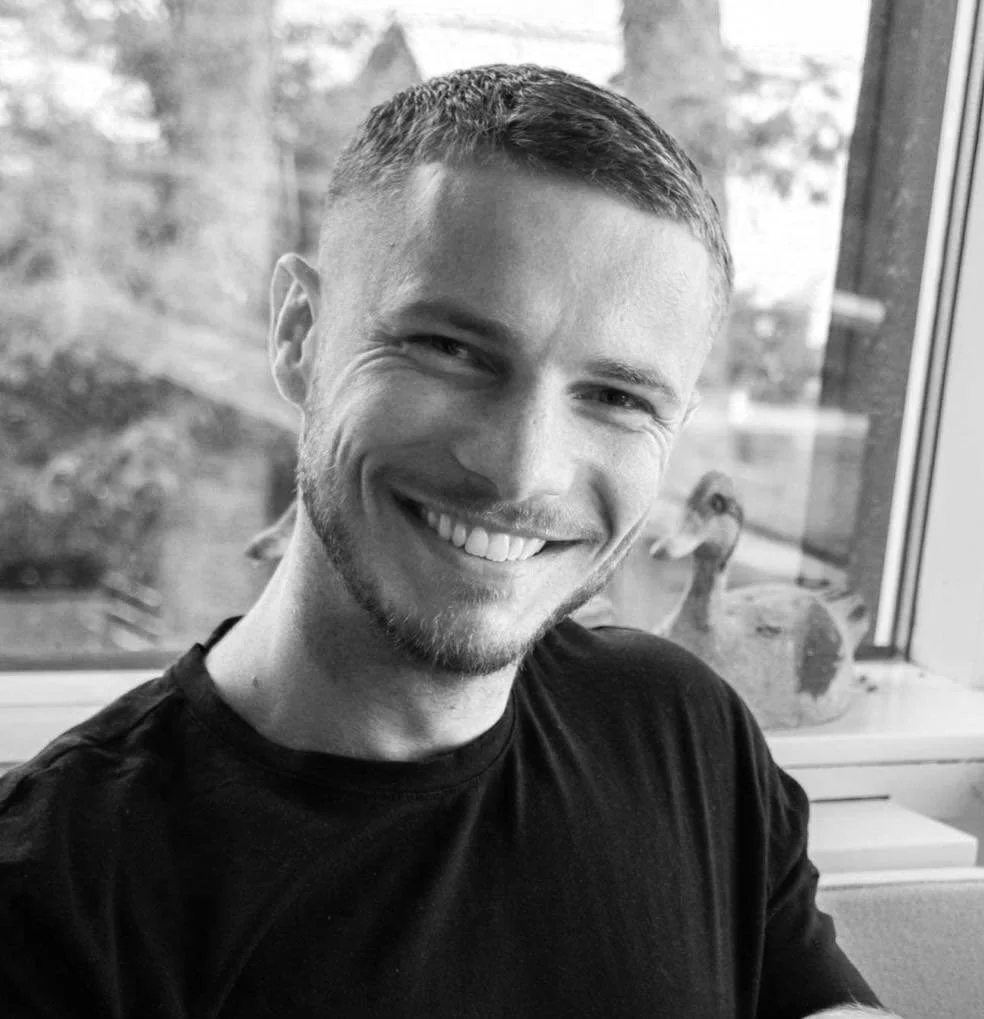 A young man with short hair and a beard smiling at the camera, sitting indoors near a window with ducks visible outside.
