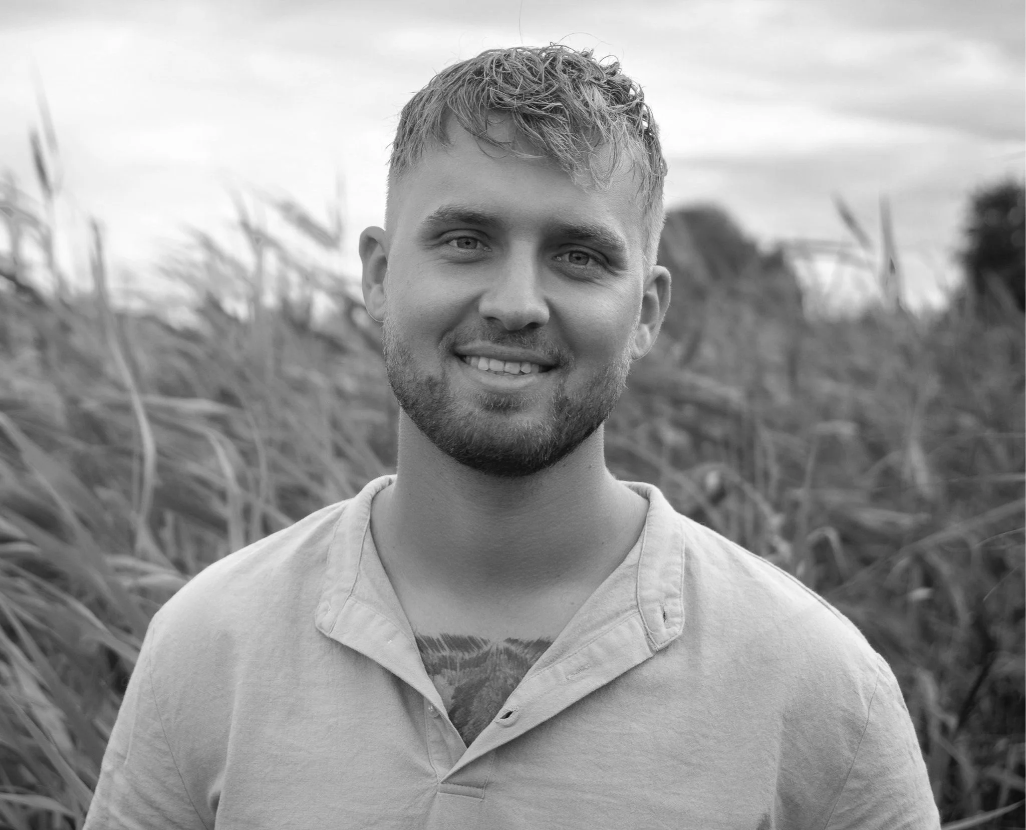 A black and white portrait of a smiling young man with short, wavy hair and a beard, standing outdoors in a field of tall grass with trees in the background.
