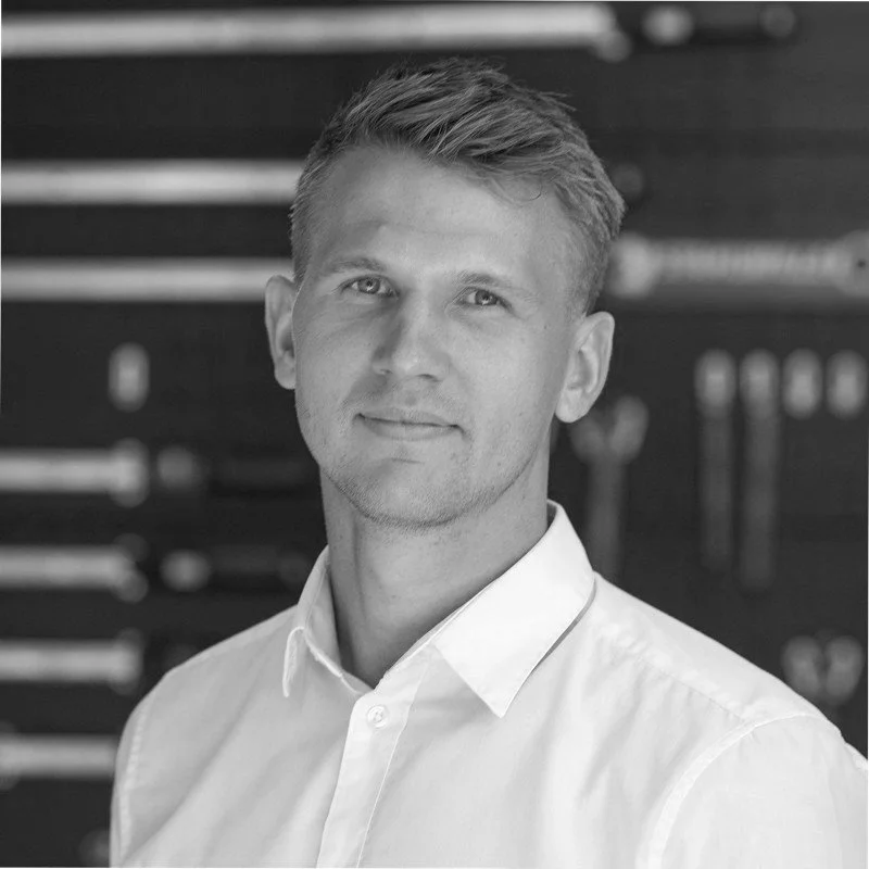 Black and white photo of a young man with short hair, wearing a white collared shirt, standing in front of a wall with tools hanging on it.