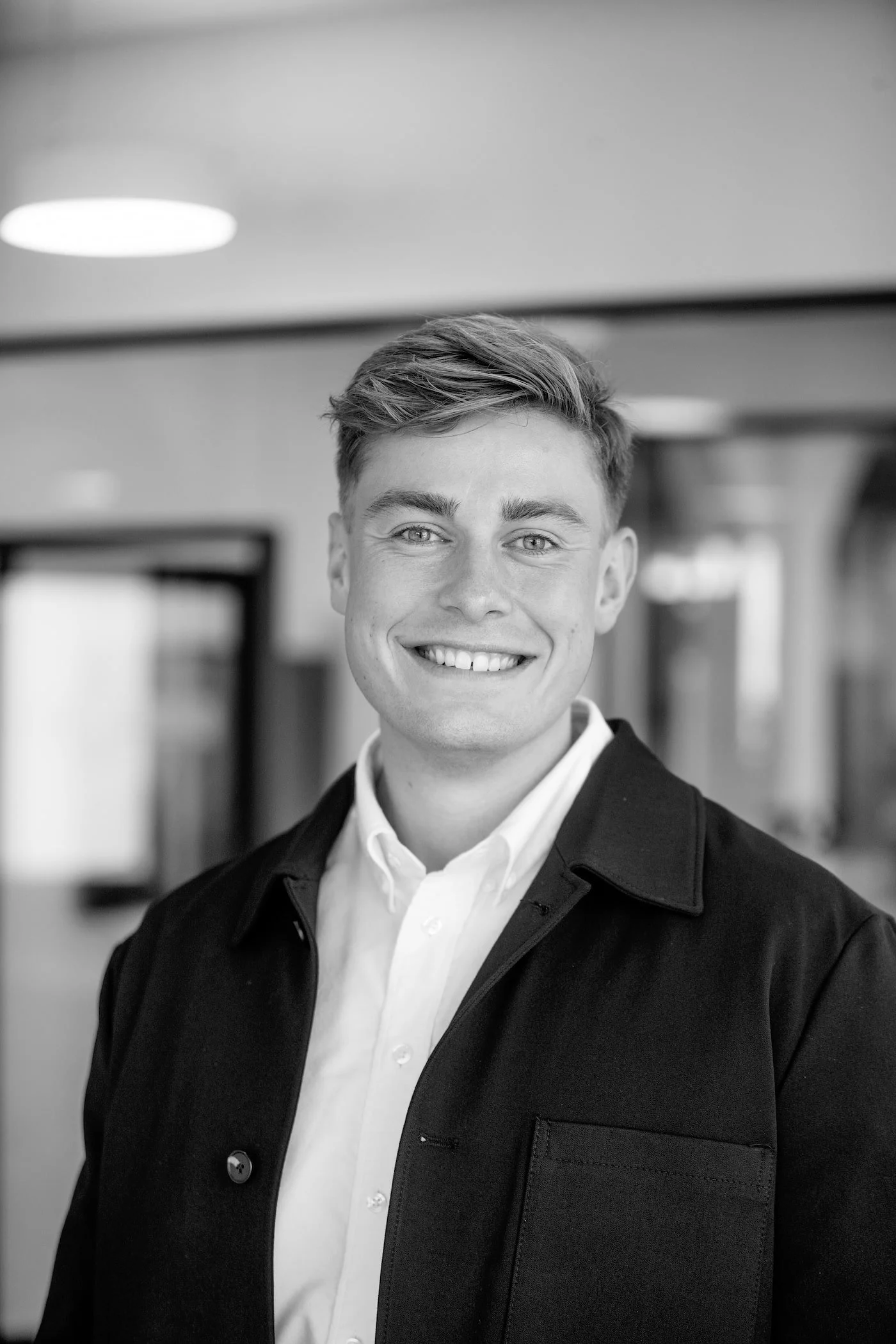 Black and white photo of a young man in a white shirt and dark jacket, smiling indoors.