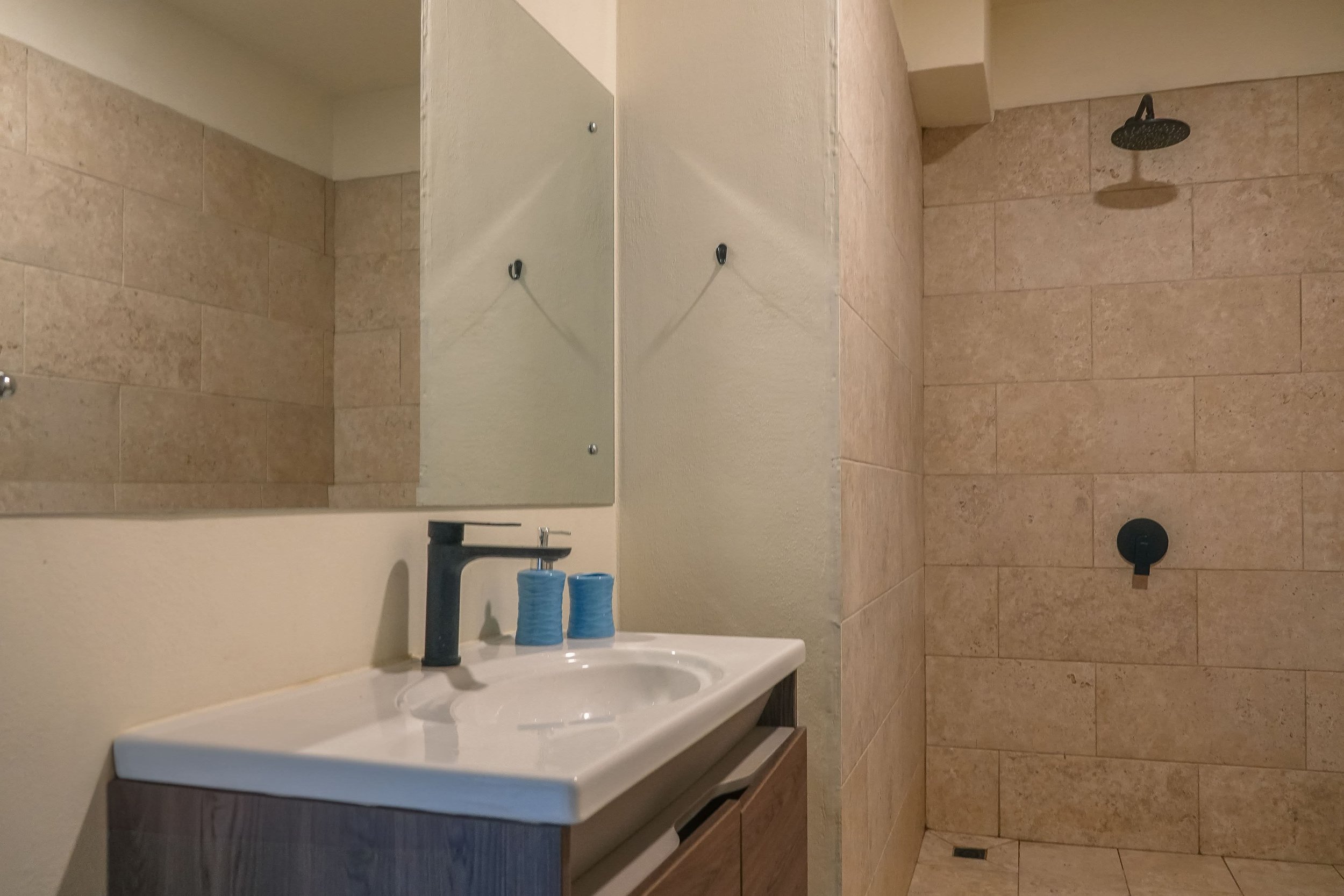 Bathroom with beige tiled walls and floor, black faucet on white sink with blue soap dispenser. Shower area with black rain showerhead, no curtain or door.