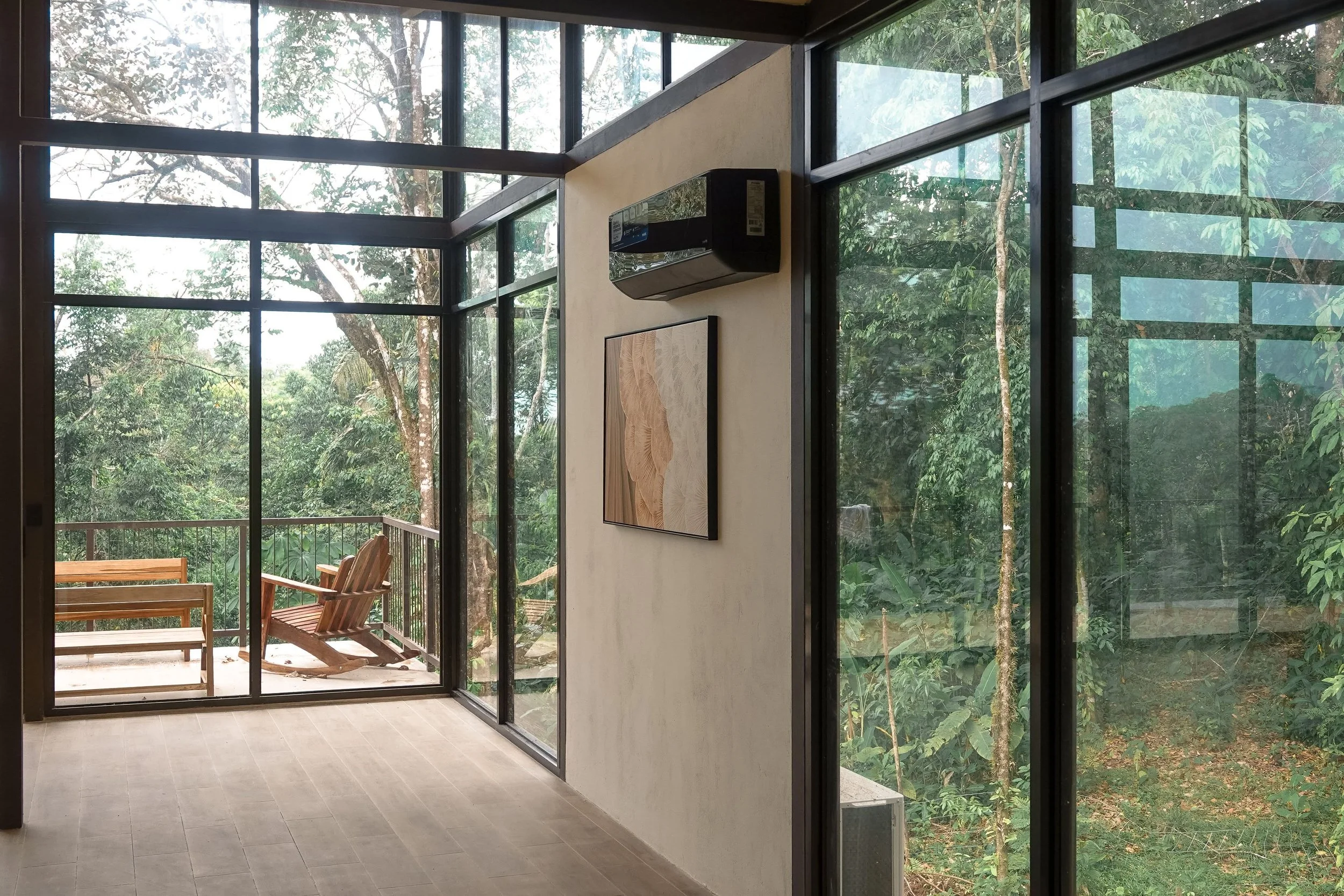 Interior view of a room with large glass walls showing trees outside, a wooden rocking chair, and a balcony with a wooden bench.