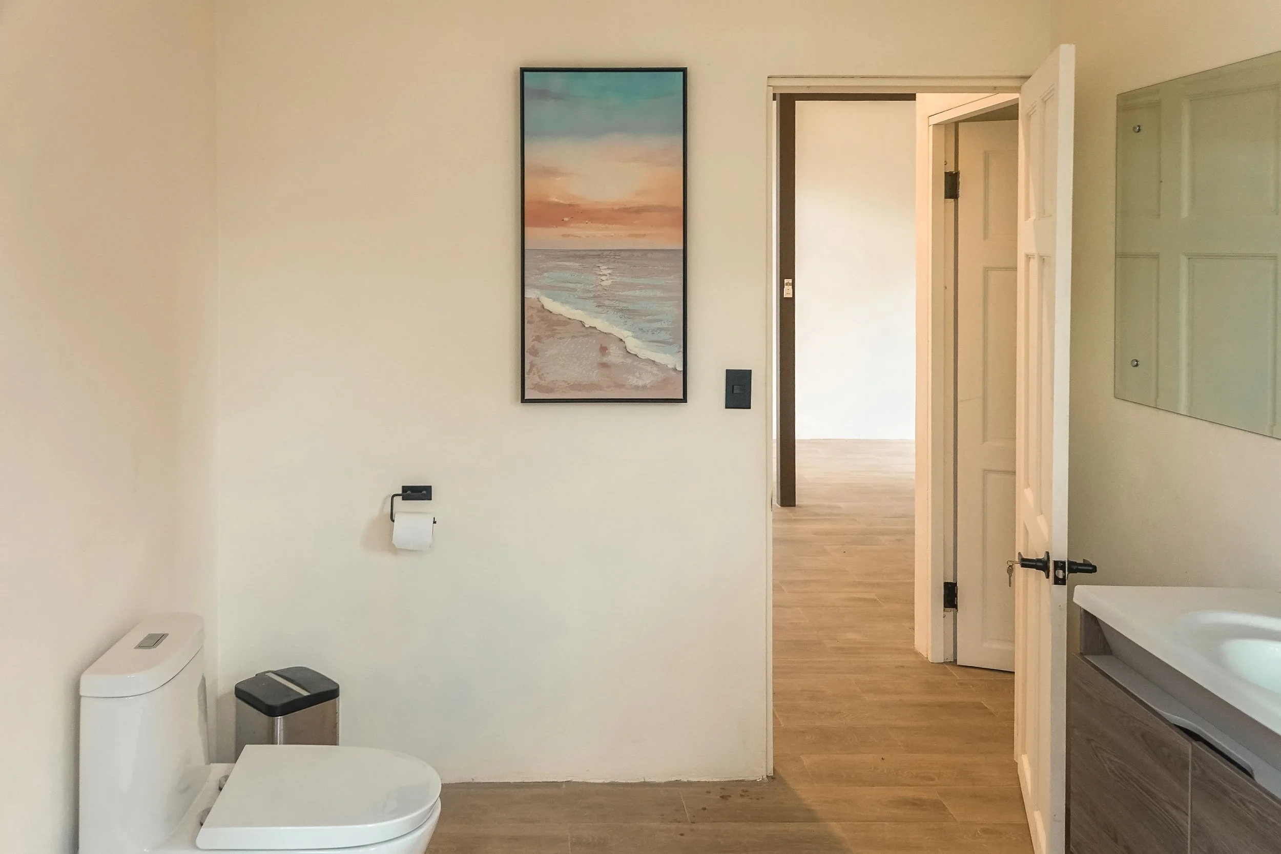 Bathroom with white walls, a toilet, a small trash bin, a painting of a beach sunset, a wall-mounted mirror, and an open door leading to a room with wooden flooring.