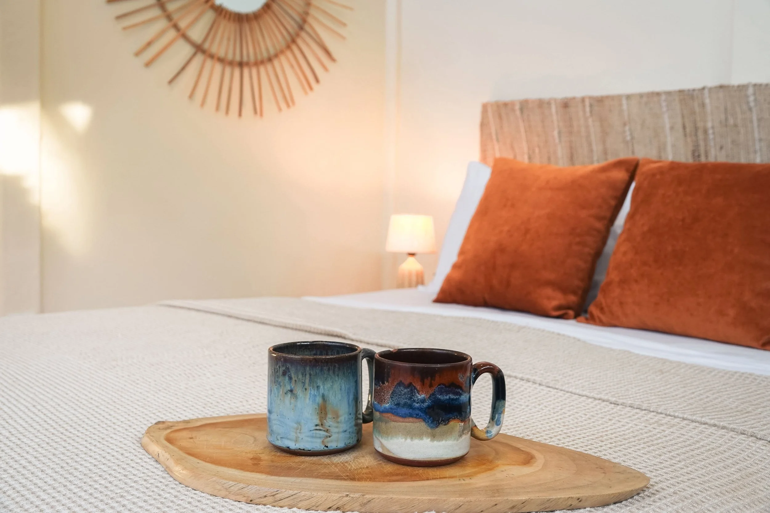 A wooden tray with two ceramic coffee mugs on a bed with beige and orange pillows, a bedside lamp, and a wall decoration in the background.