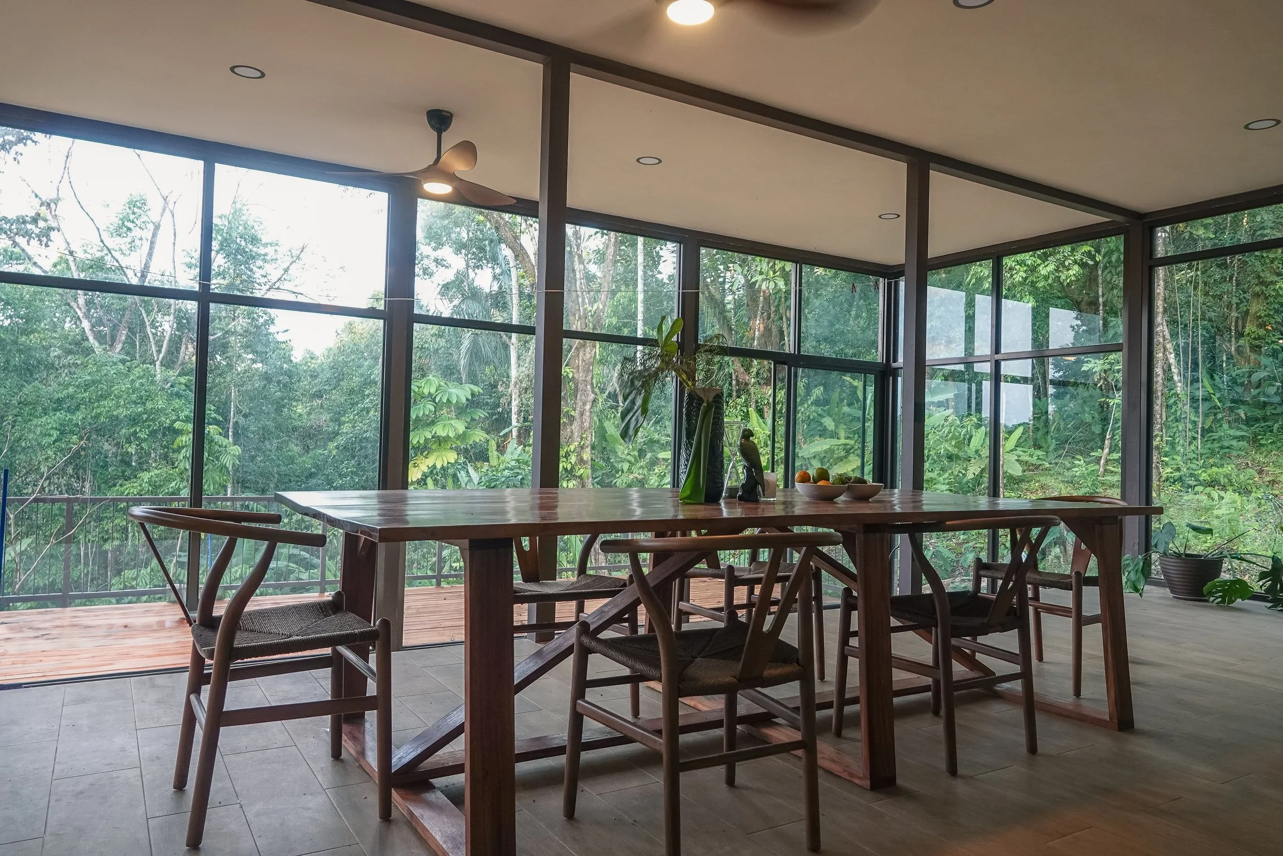 Modern dining area with wooden table and chairs, large glass windows overlooking lush green forest, with ceiling fans and plants inside.