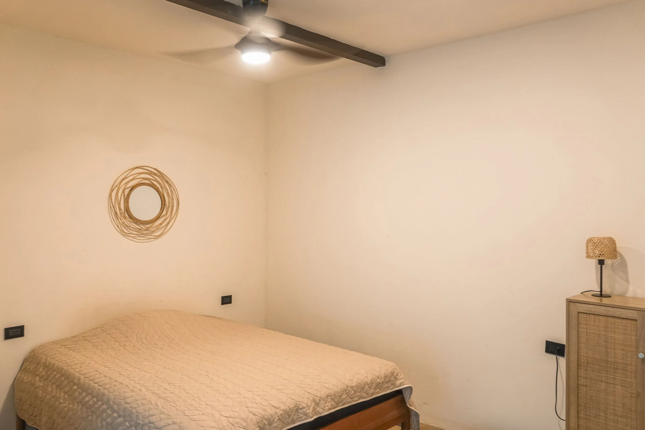 A minimalist bedroom with a bed covered in a beige quilt, a round wicker mirror on the wall above the bed, a ceiling fan with a light, a wooden dresser with a wicker lamp, and multiple black electrical outlets on the wall.