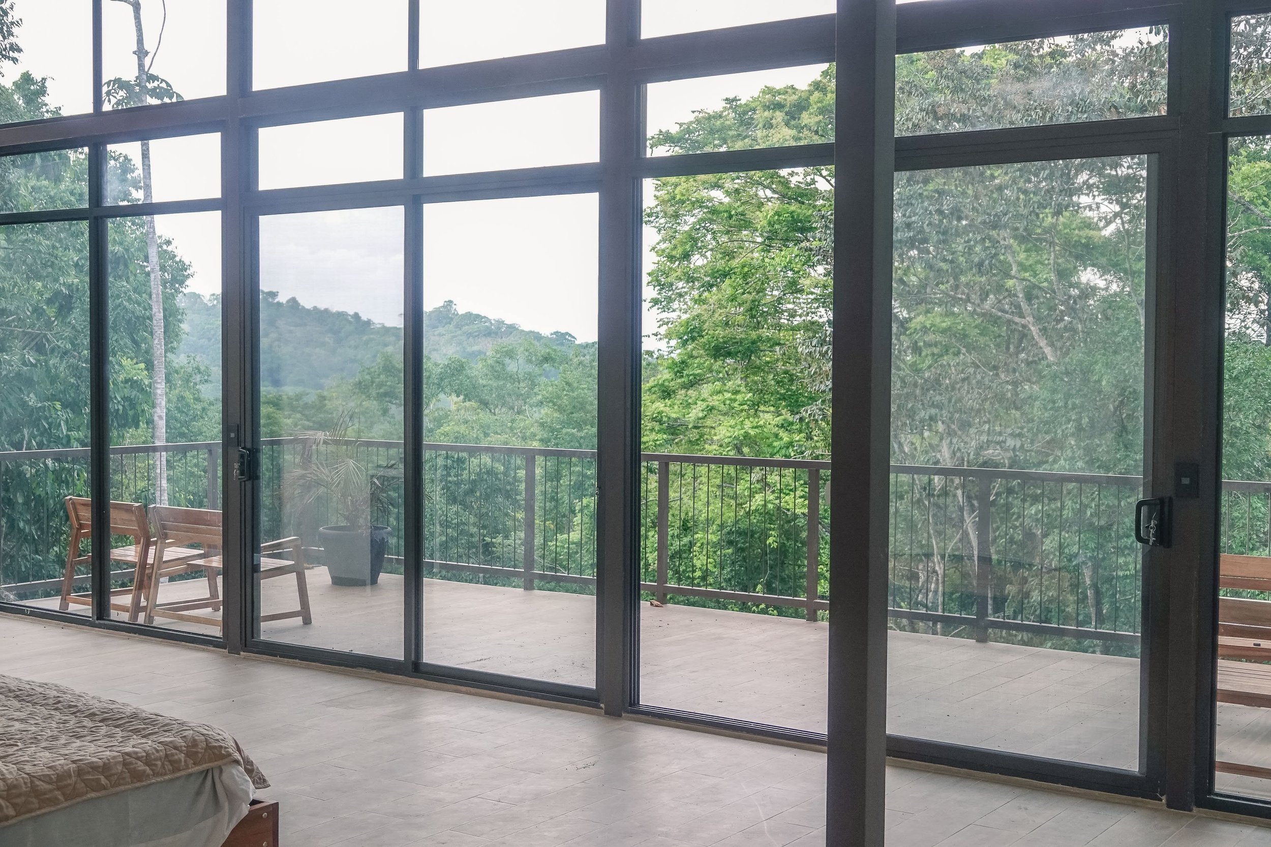 View through large glass sliding doors onto a balcony with a bench and potted plant, overlooking a lush green forested area.