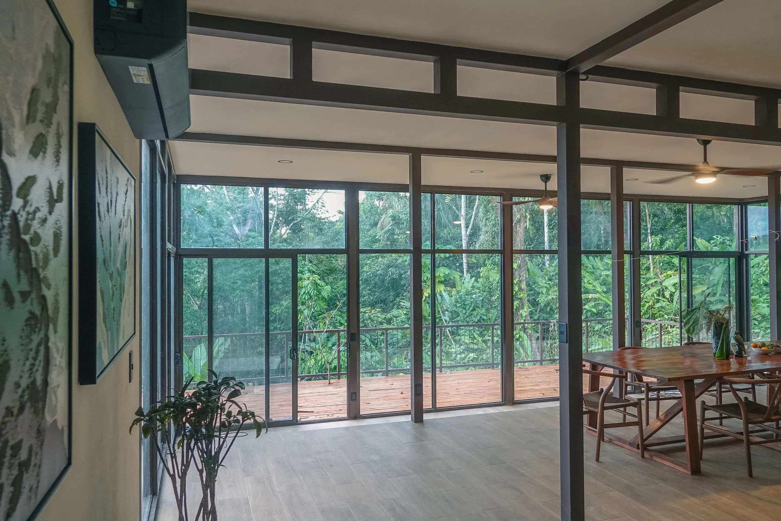 Modern living space with large floor-to-ceiling windows overlooking a lush green forest, wooden deck outside, dining table with chairs, indoor plants, ceiling fans, and mounted air conditioning.