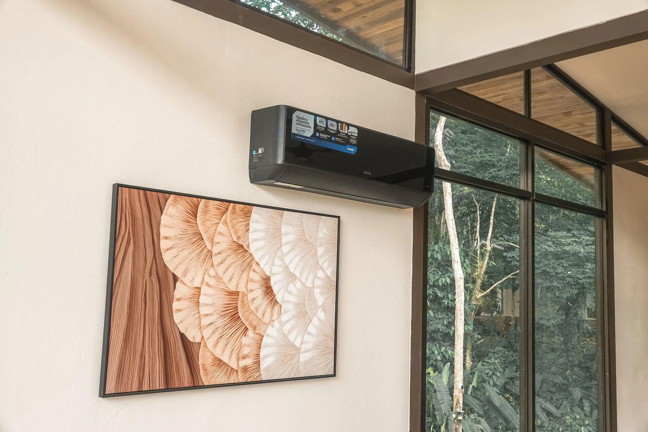 Wall-mounted black air conditioning unit above a framed picture of earth-toned shell patterns on a white wall, adjacent to a large glass window showing greenery outside.