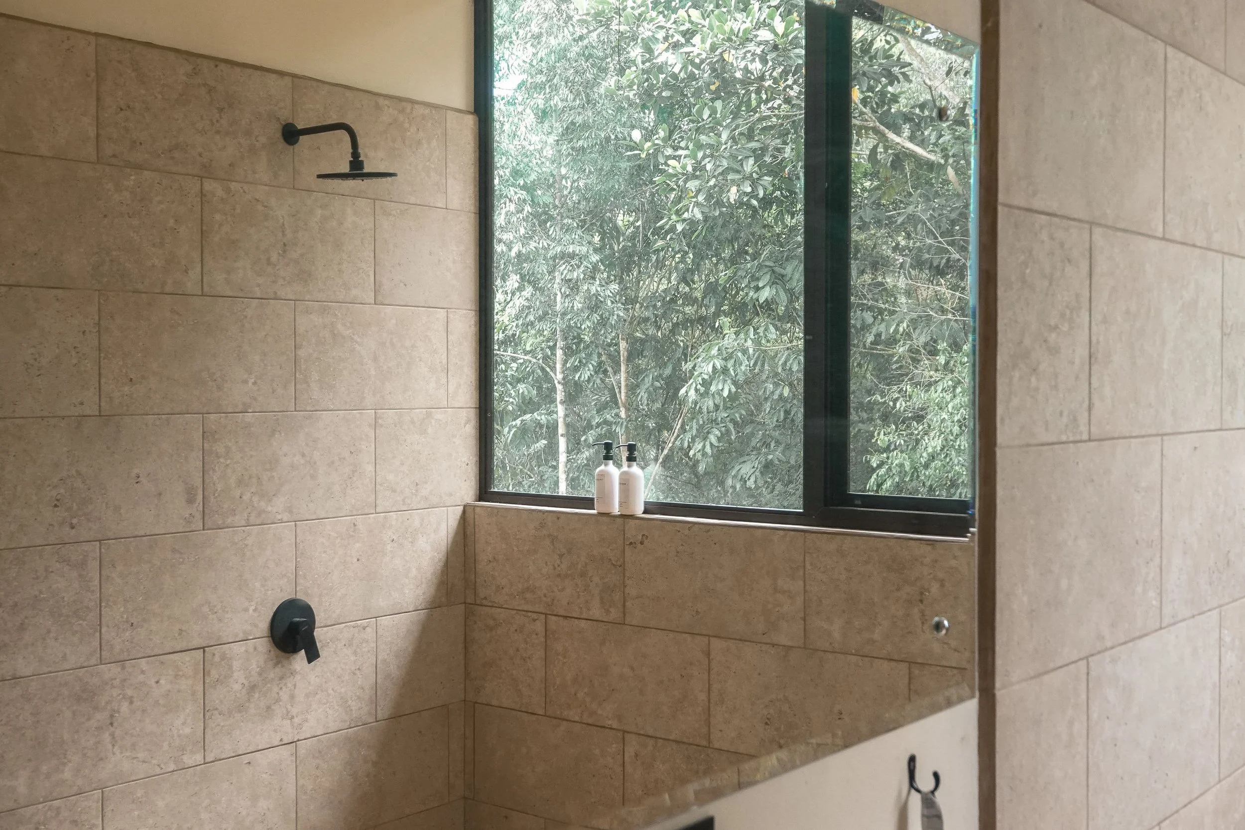 Shower area with a black showerhead, a black control handle, beige tiled walls, a large window showing trees outside, and two soap bottles on the windowsill.