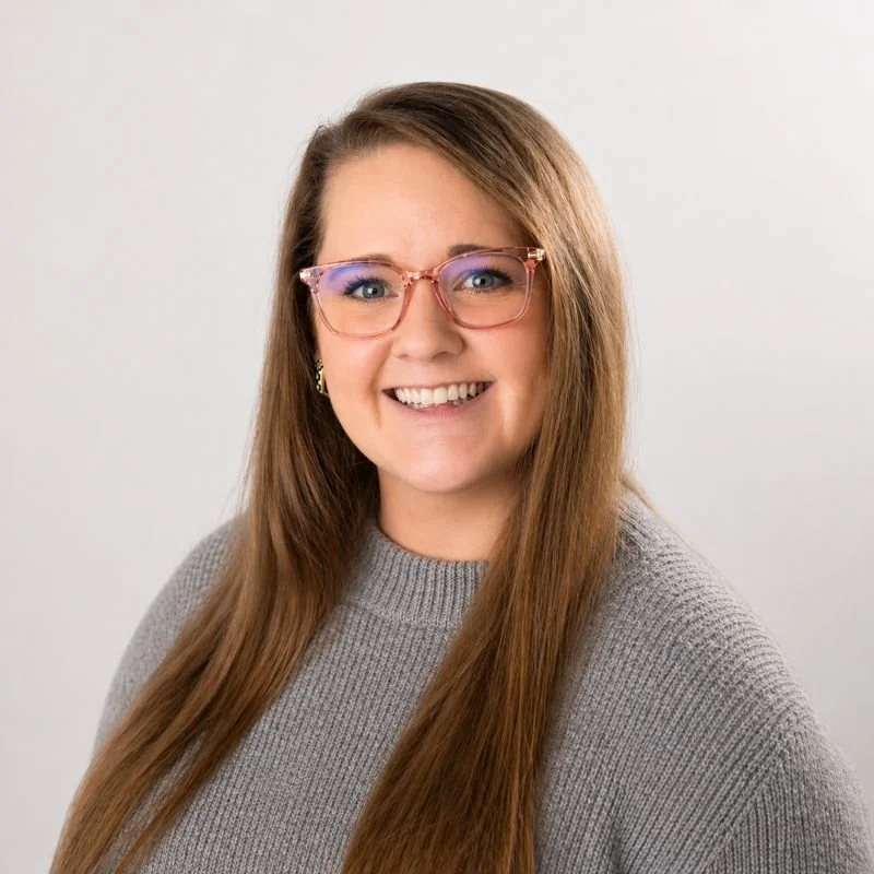 A woman with long brown hair, wearing pink glasses and a gray sweater, smiling at the camera against a plain light gray background.