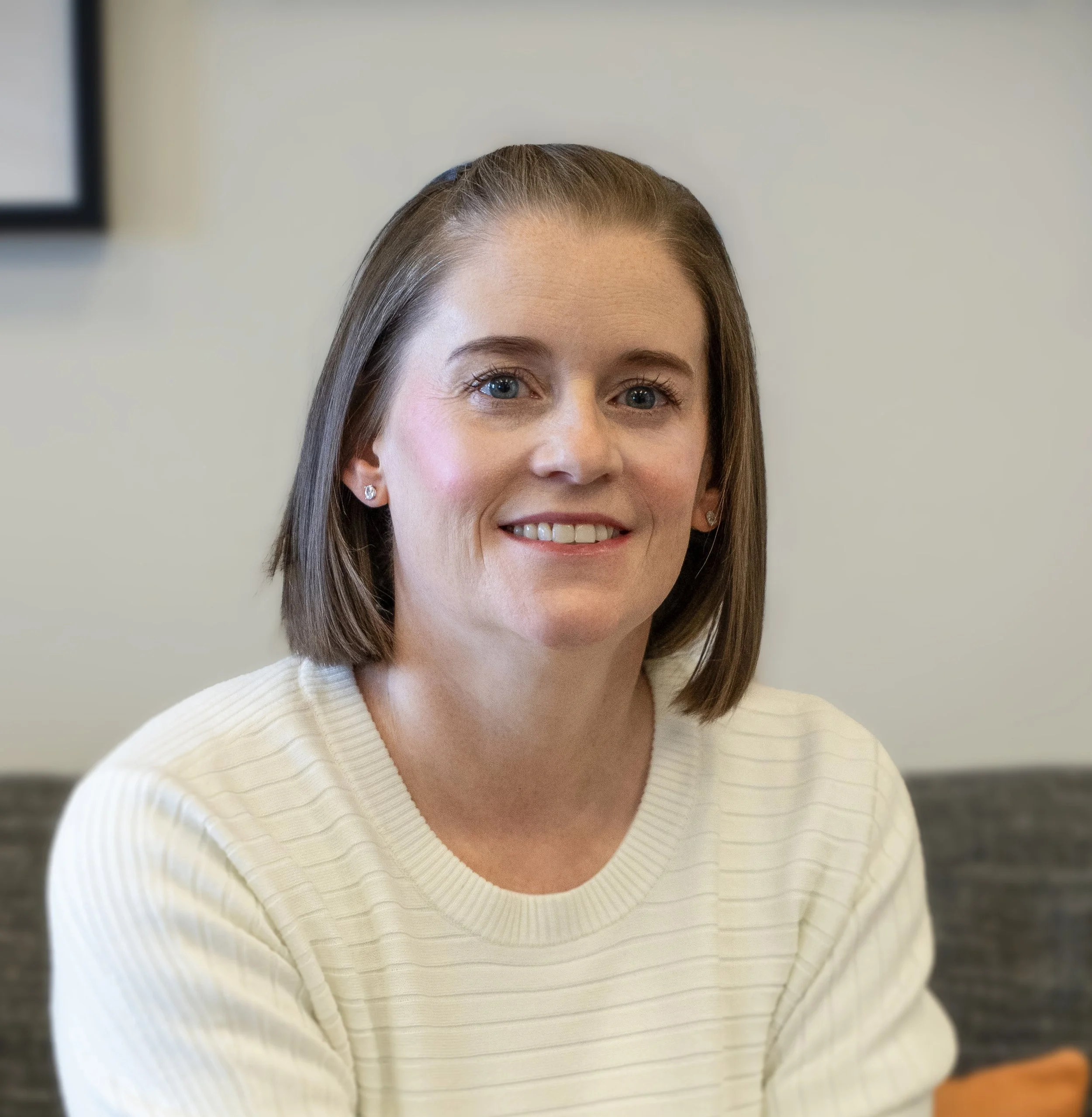 Close-up of a woman with short brown hair, blue eyes, wearing diamond earrings and a cream-colored ribbed sweater. The background is plain and out of focus.
