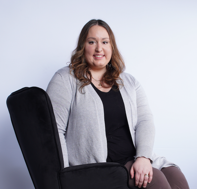 Woman with brown hair sitting on a black chair, smiling at the camera, wearing a gray cardigan over a black shirt, against a plain white background.