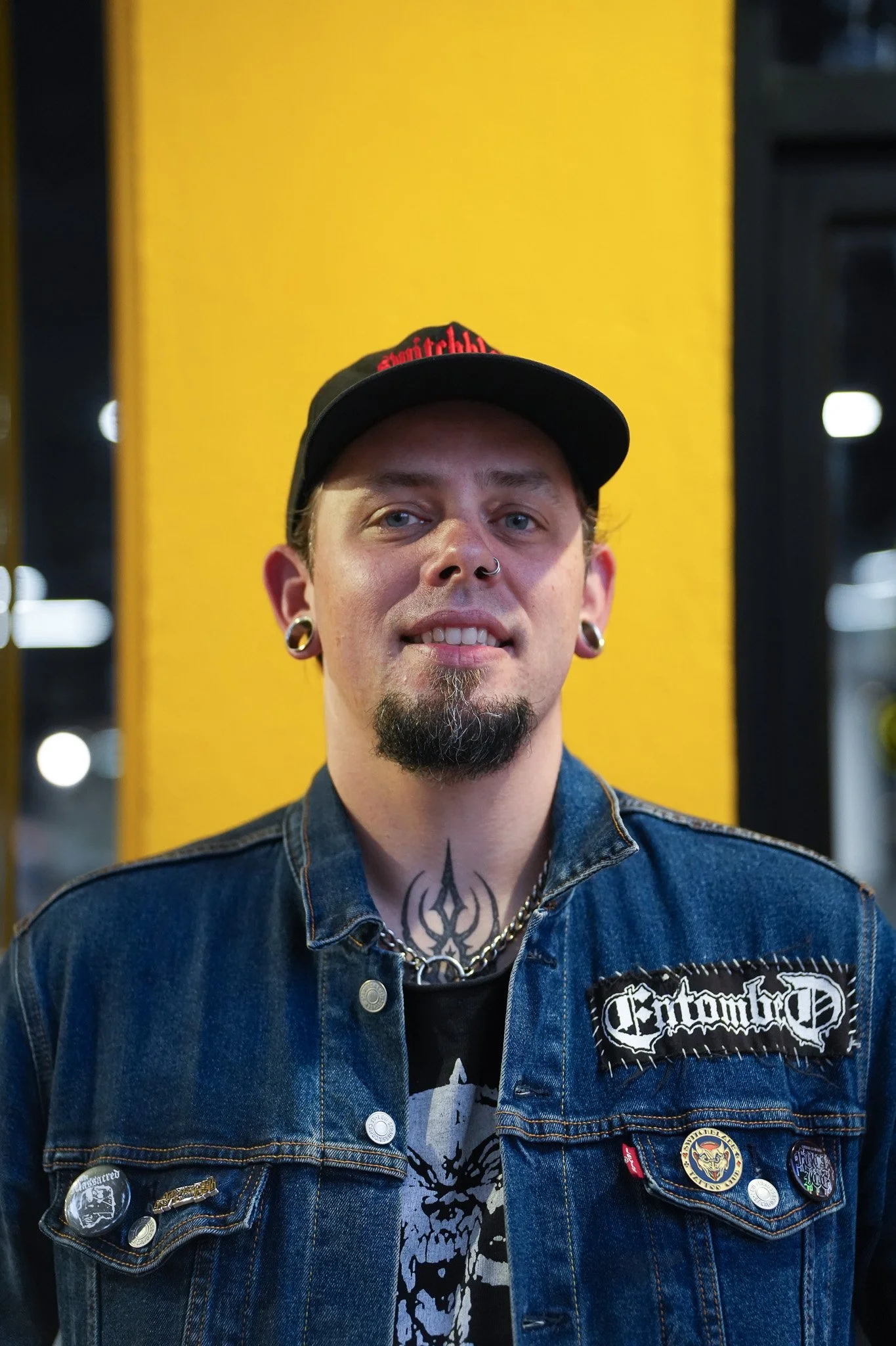 Close-up of a young man with piercings and tattoos wearing a denim jacket with various patches and buttons, standing in front of a yellow background inside a store or building.