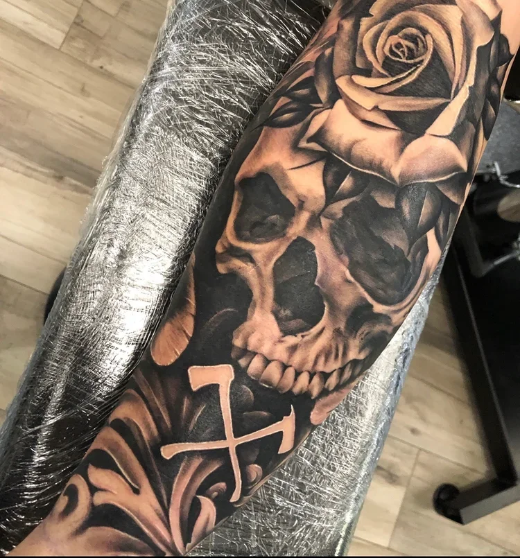 Tattoo of a skull with roses and leaves, along with a cross symbol on the forearm.