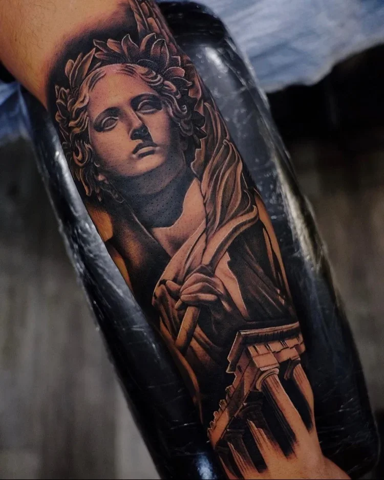 A tattoo of a classical style bust of a woman with a crown of laurel leaves, resembling a Greek or Roman sculpture, on a person's arm.