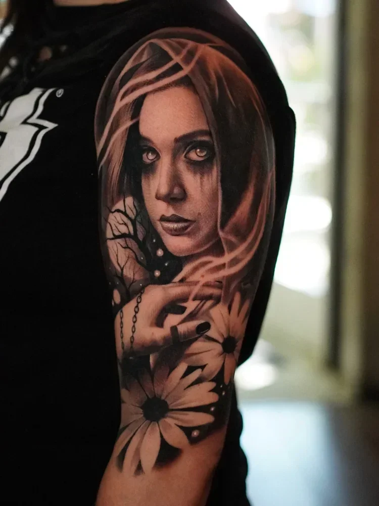Tattoo of a woman's face with dark makeup, surrounded by flowers and branches, on a person's upper arm.