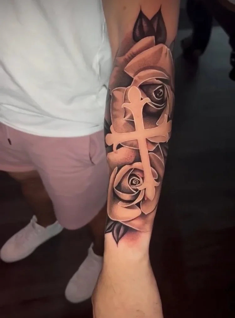 Tattoo of a cross and roses on a person's forearm.