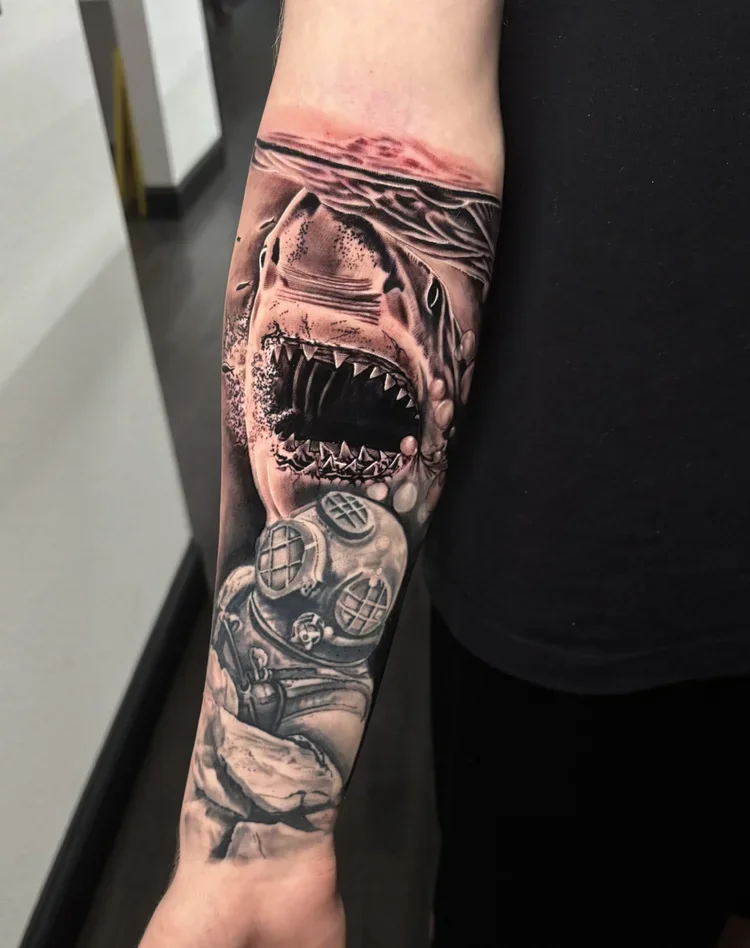 Black and gray tattoo of a great white shark attacking a diver with scuba gear, on a person's forearm.
