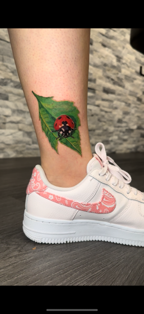 Tattoo of a ladybug on a green leaf on a person's ankle, wearing a white sneaker with pink designs.
