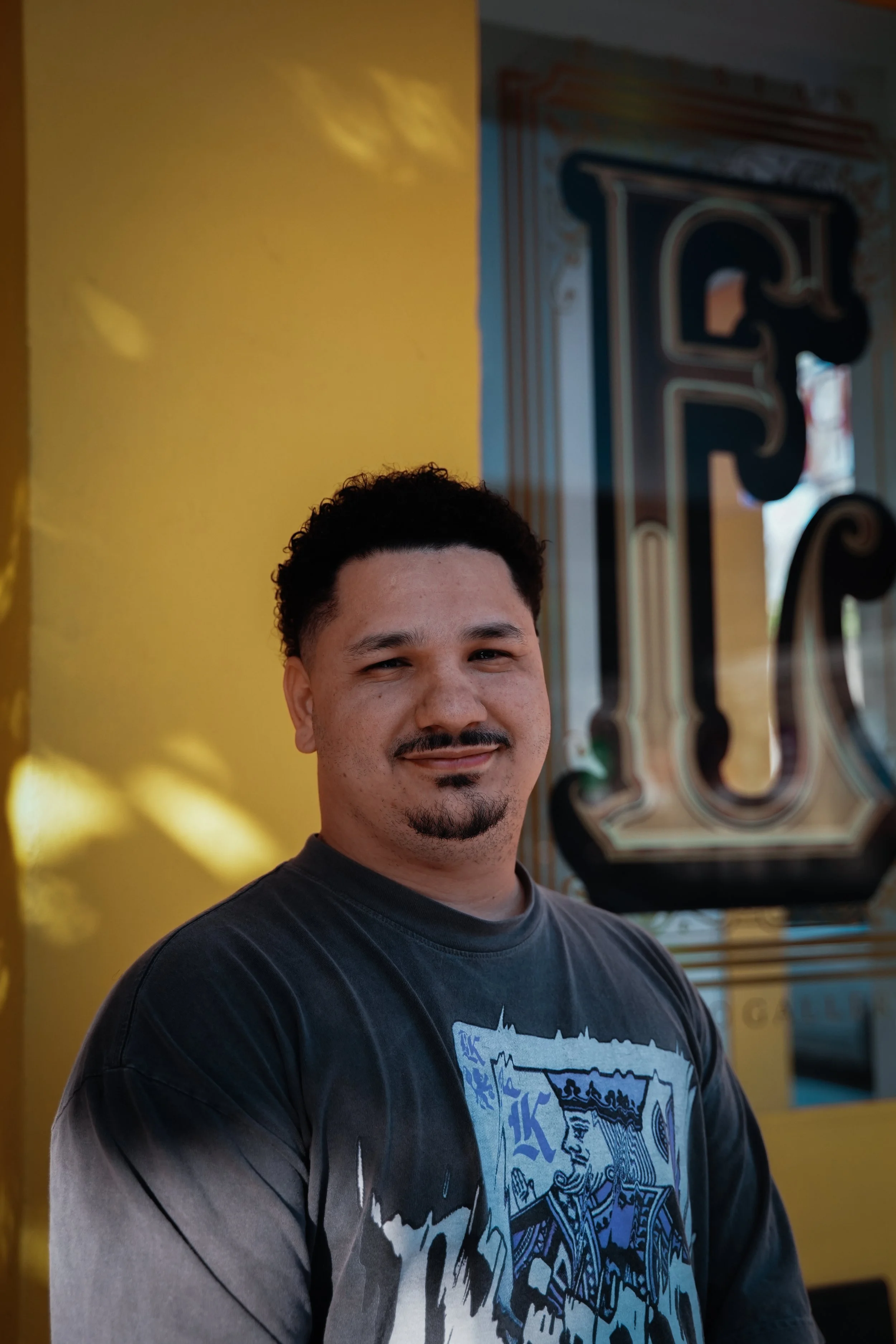 A smiling man with dark curly hair and a goatee stands in front of a yellow wall and a colorful sign with a large letter 'F'.