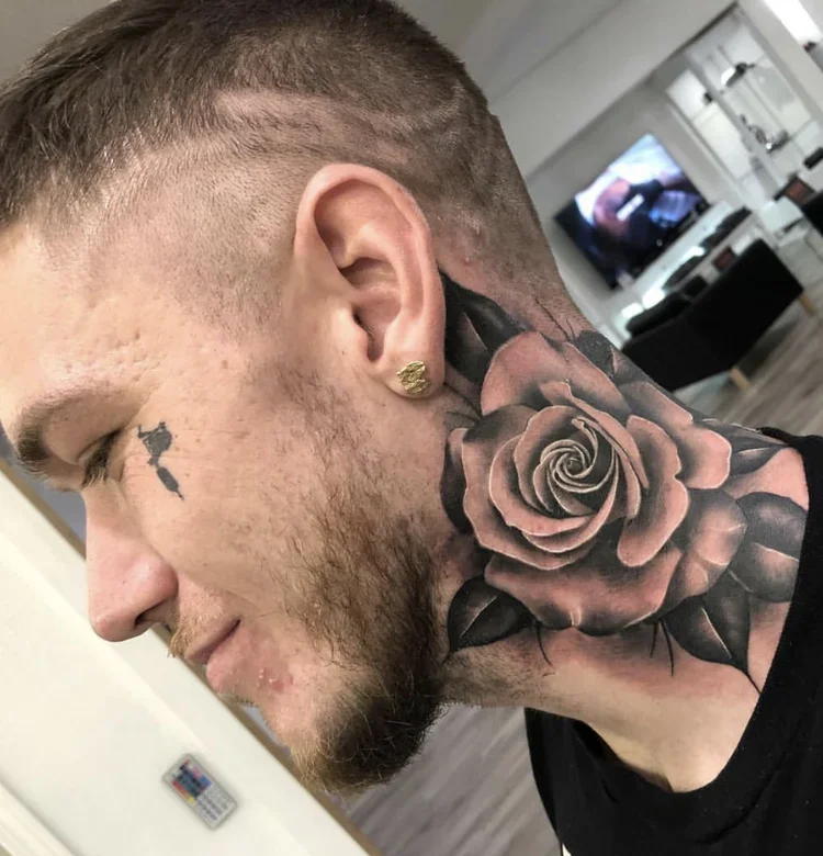 Profile of a young man with short hair and beard, large black and gray rose tattoo on neck, wearing a gold earring, in a modern room.
