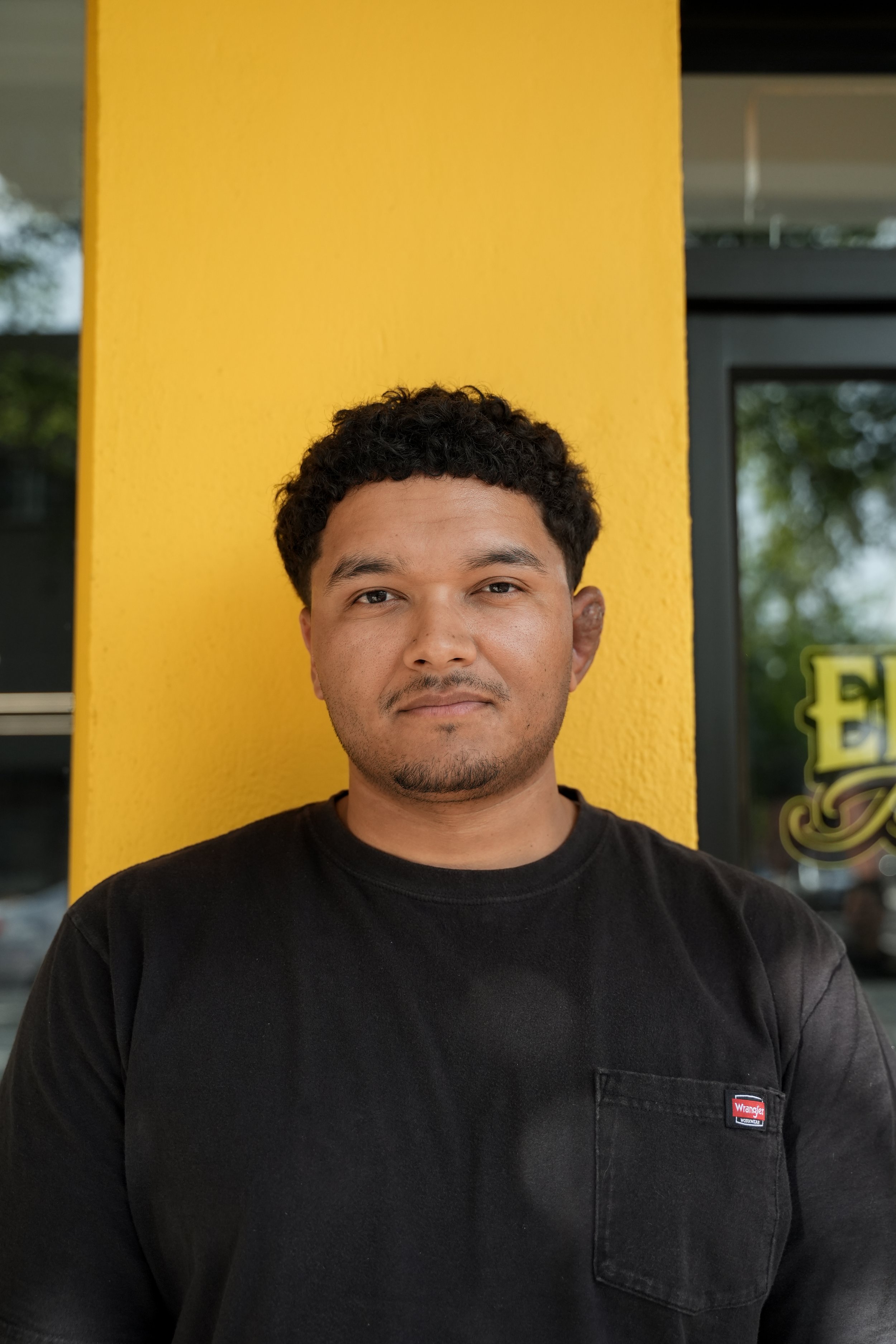 A man with dark, curly hair and a slight beard, wearing a black T-shirt, standing against a bright yellow wall with a window and some greenery reflected in the background.