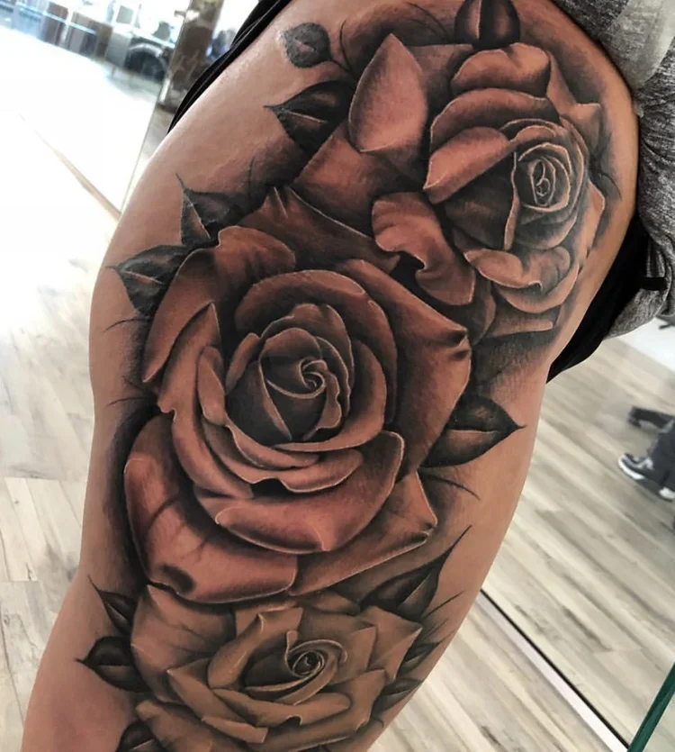 Detailed black and gray tattoo of three large roses with leaves on a person's thigh.