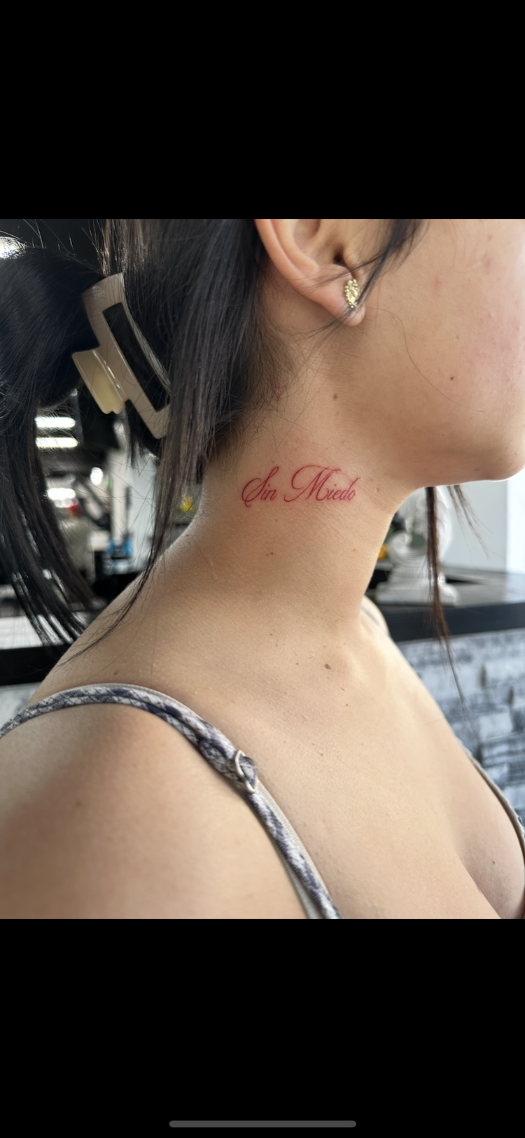 Close-up of a woman's neck tattooed with the words 'Sin Miedo' in red cursive, with part of her face, ear with earrings, and ponytail visible.