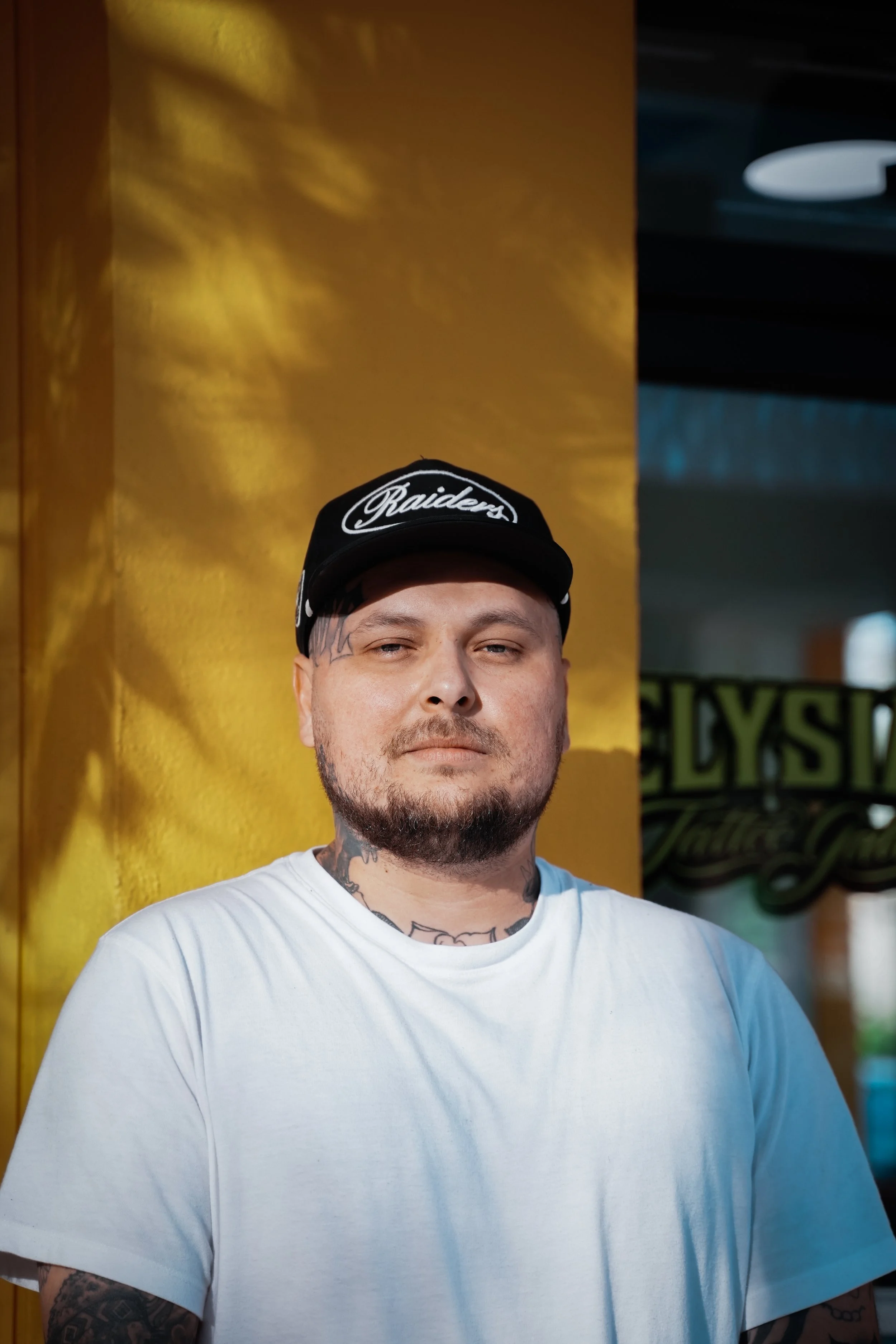 Portrait of a man with tattoos wearing a white t-shirt and a black Raiders cap standing in front of a yellow wall.