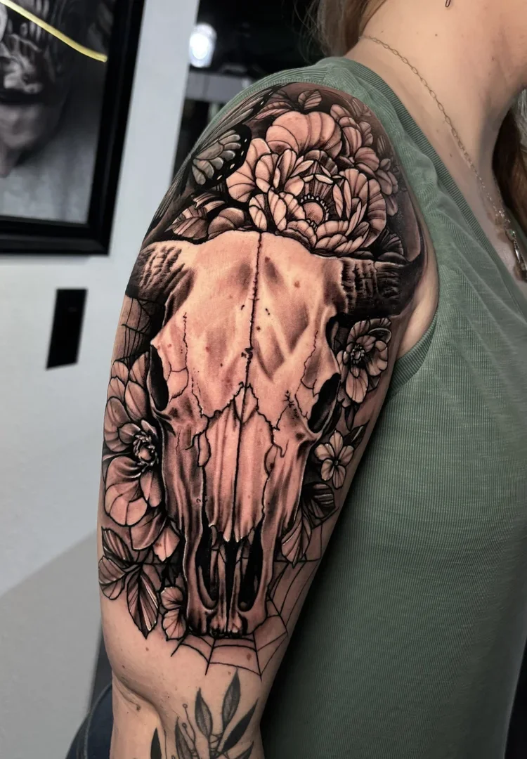 Tattoo of a animal skull surrounded by flowers on a person's upper arm.