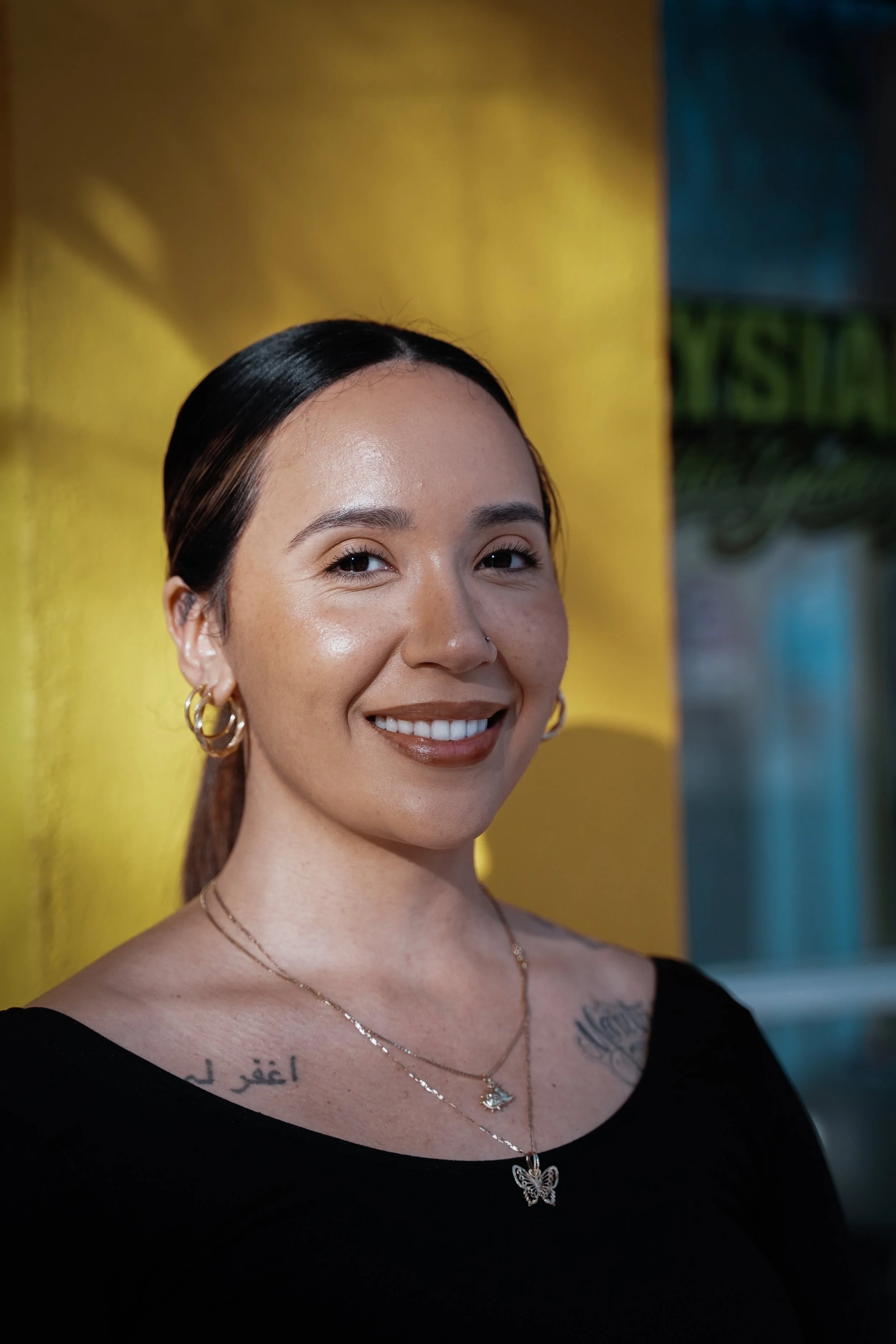 A smiling woman with dark hair tied back, wearing gold hoop earrings and layered necklaces, standing in front of a vibrant yellow wall.
