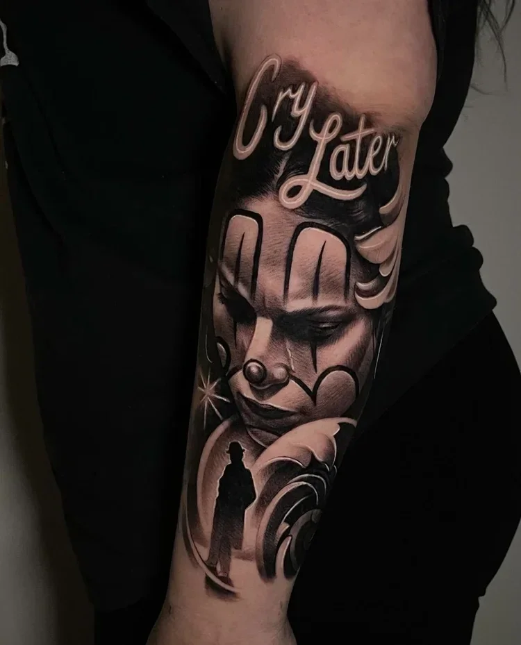 Black and gray realistic tattoo of a child clown face with makeup, a small nose, and a sad expression, with the words "Cry Later" above and a silhouette of a person standing under the face, with swirling patterns and stars adding artistic detail.