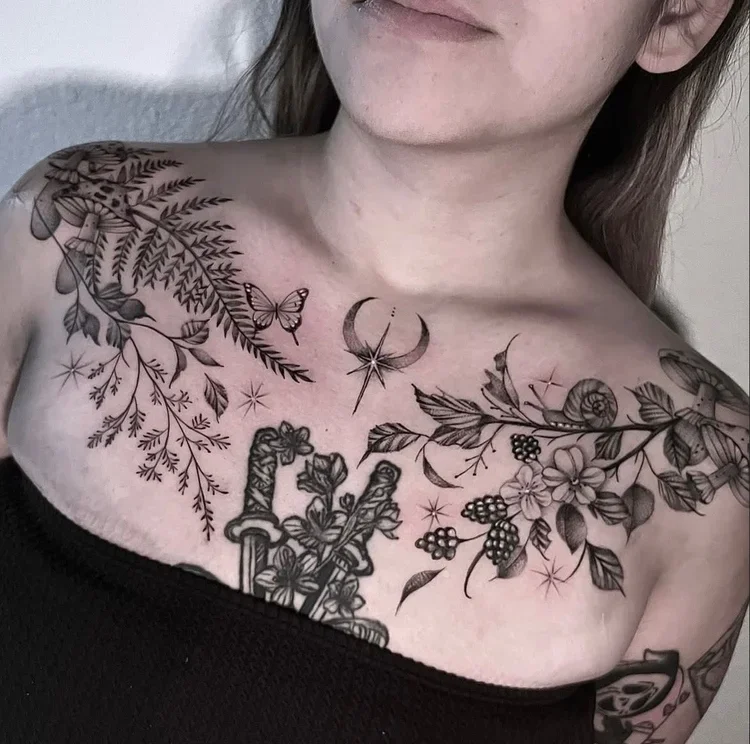 Close-up of a person's chest tattoo featuring botanical elements like leaves, flowers, and berries, along with symbols such as a crescent moon, a star, and two swords.
