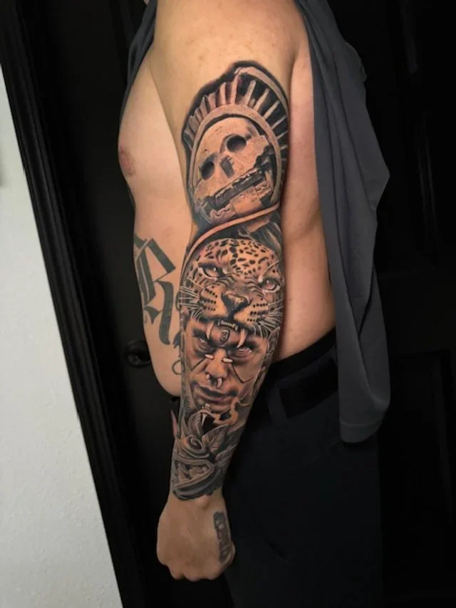 Tattoo of a skull with a helmet and a jaguar face, with a roaring human face below, on a person's upper arm.