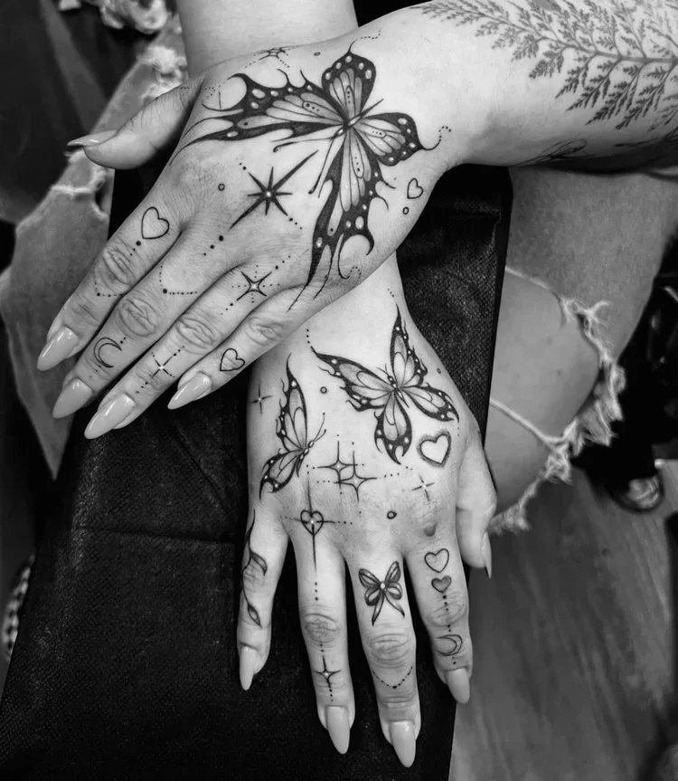 Black and white tattoo of butterflies with hearts and star-like designs on hands and nearby arm.