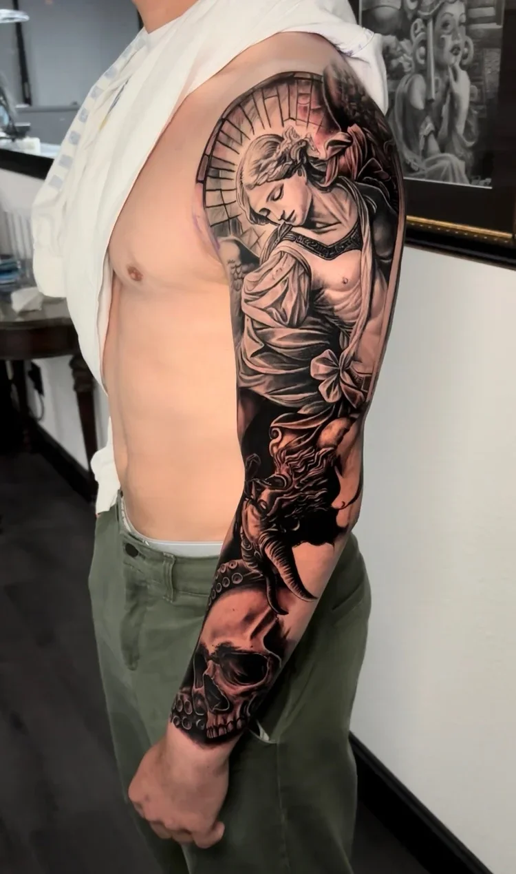 Black and gray sleeve tattoo featuring a woman, angel wings, an elephant, a skull, and decorative elements.