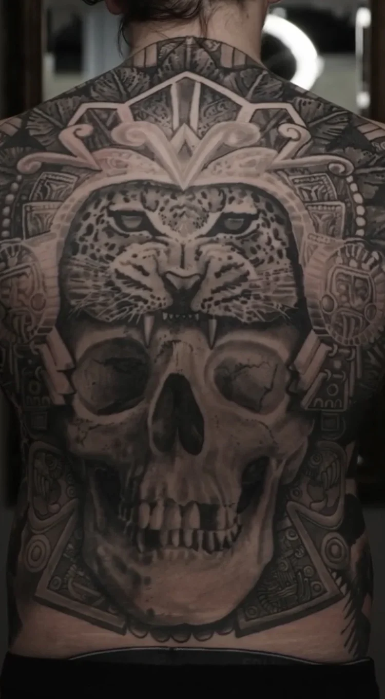 Tattoo of a leopard's face with piercing eyes, partially inside a skull, surrounded by intricate patterns and geometric designs on a person's back.