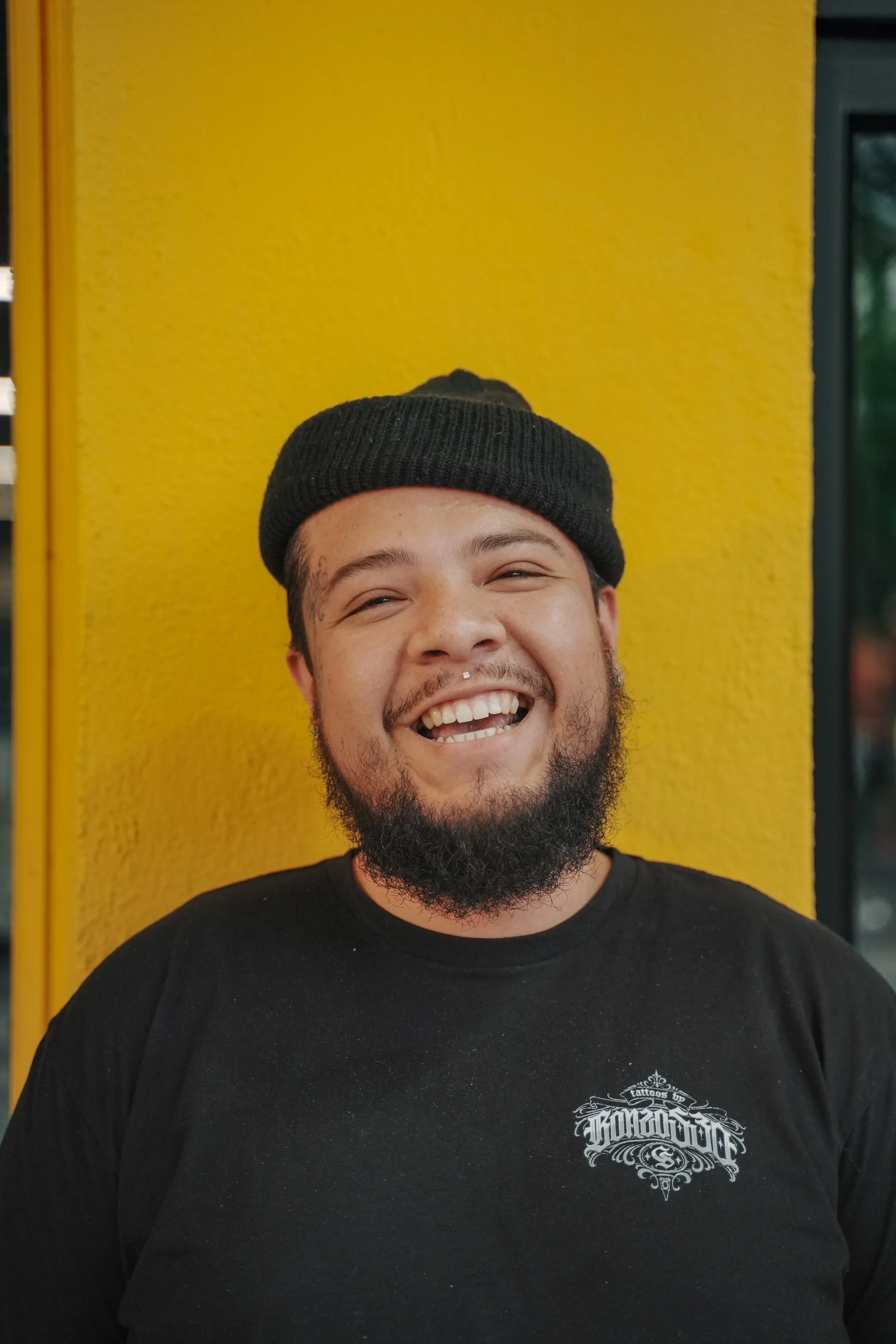 A smiling man with a beard and mustache wearing a black beanie and black t-shirt, standing in front of a yellow wall.