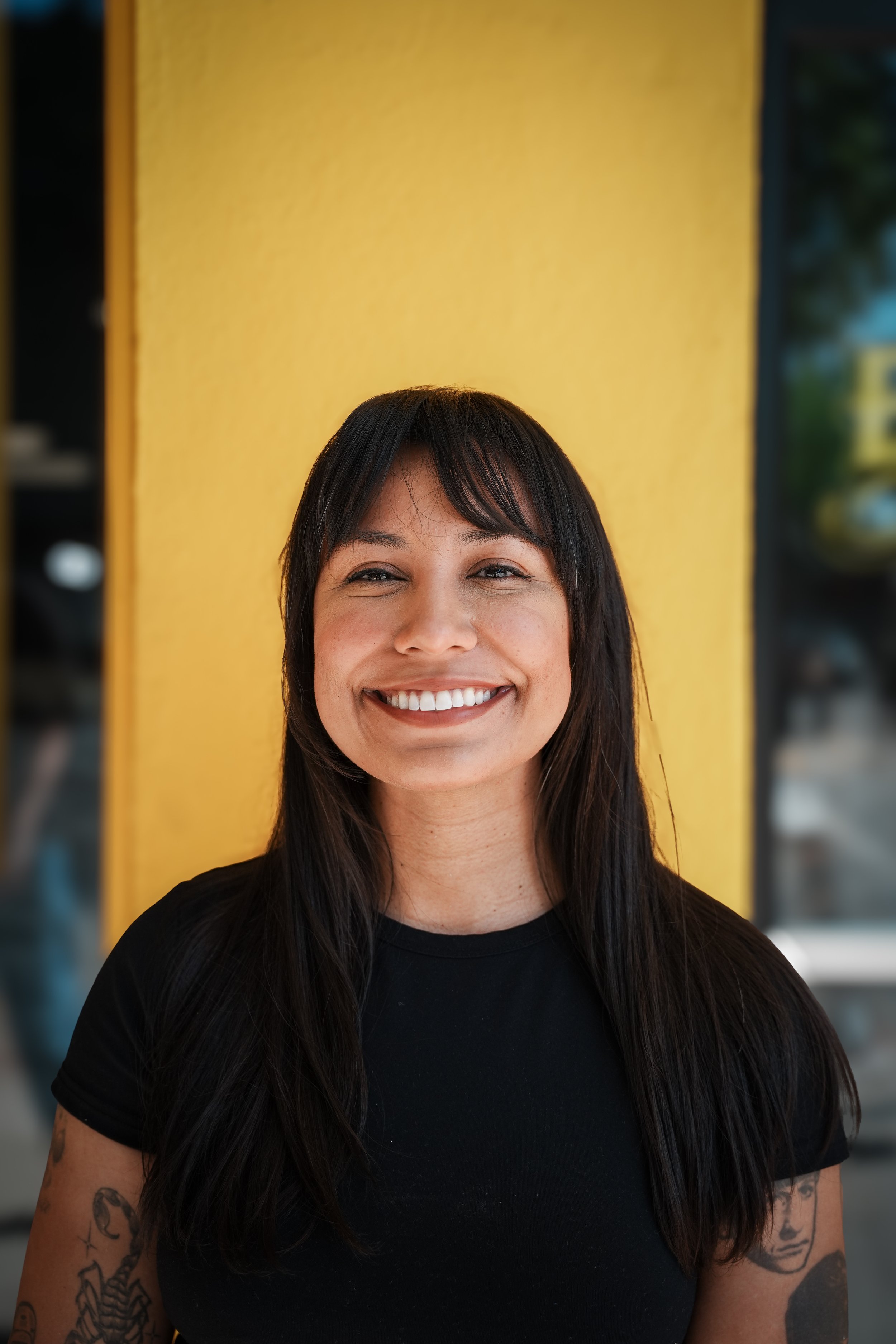 A woman with long dark hair and tattoos smiling in front of a yellow wall.