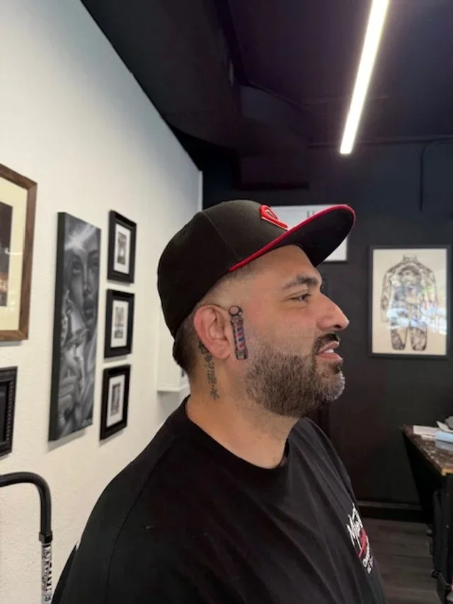 A man with a beard and tattoos, wearing a black cap with a red border and black shirt, standing in an art gallery with black and white framed artwork on the walls.