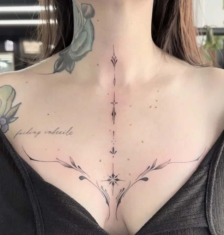 Close-up of a woman's chest with a vertical symmetrical tattoo design spanning from the collarbone down between her breasts, and a tattoo on her left shoulder that says "fucking imbecile."