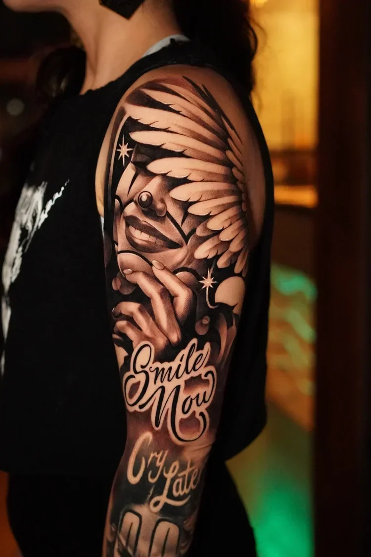 A black and gray realistic tattoo on a person's upper arm featuring a smiling woman with closed eyes, holding her chin with one hand. The woman has wings on her head, stars, and a moon. The tattoo includes the words "Smile Now" and "Cry Later" in sty