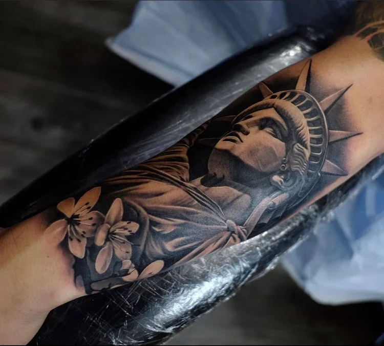 Close-up of a tattoo on someone's arm featuring the Statue of Liberty's face with flowers surrounding it.