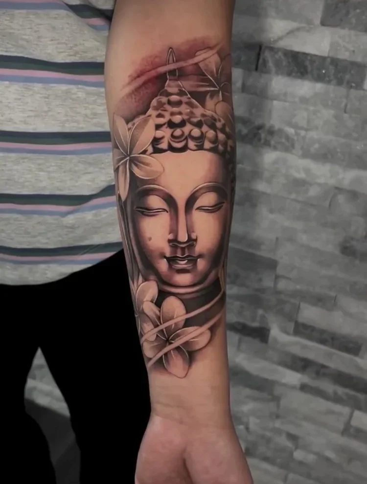 Tattoo of a serene Buddha face with closed eyes, surrounded by flowers and intricate patterns on a person's arm.