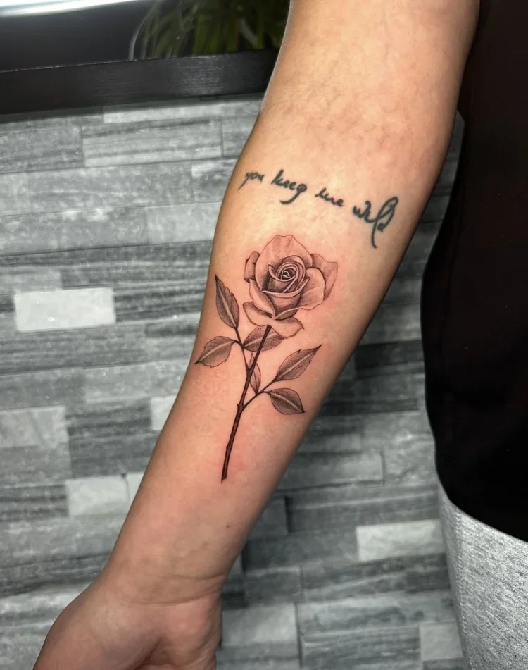 A person's forearm tattoo with a realistic rose and a quote above it that reads 'you hug me' in cursive.