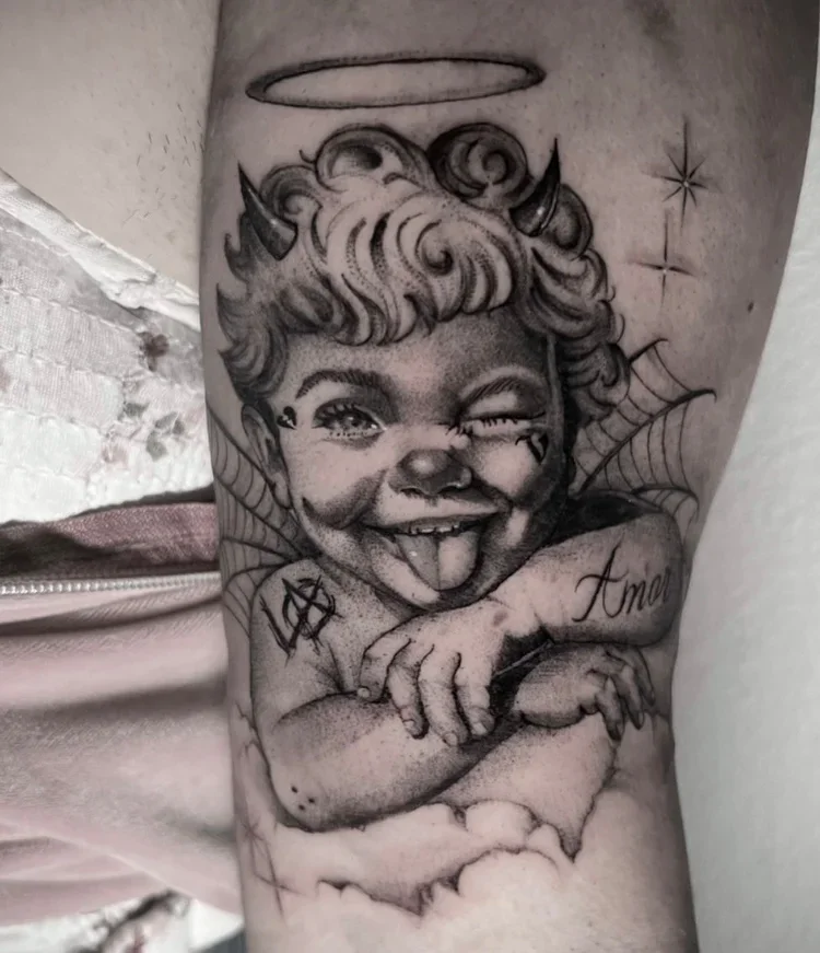 Tattoo of a mischievous child with devil horns, a halo above, and spiderwebs in the background, sticking out tongue and winking.